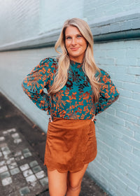 Woman wearing a floral blouse and brown skirt against a light blue wall.