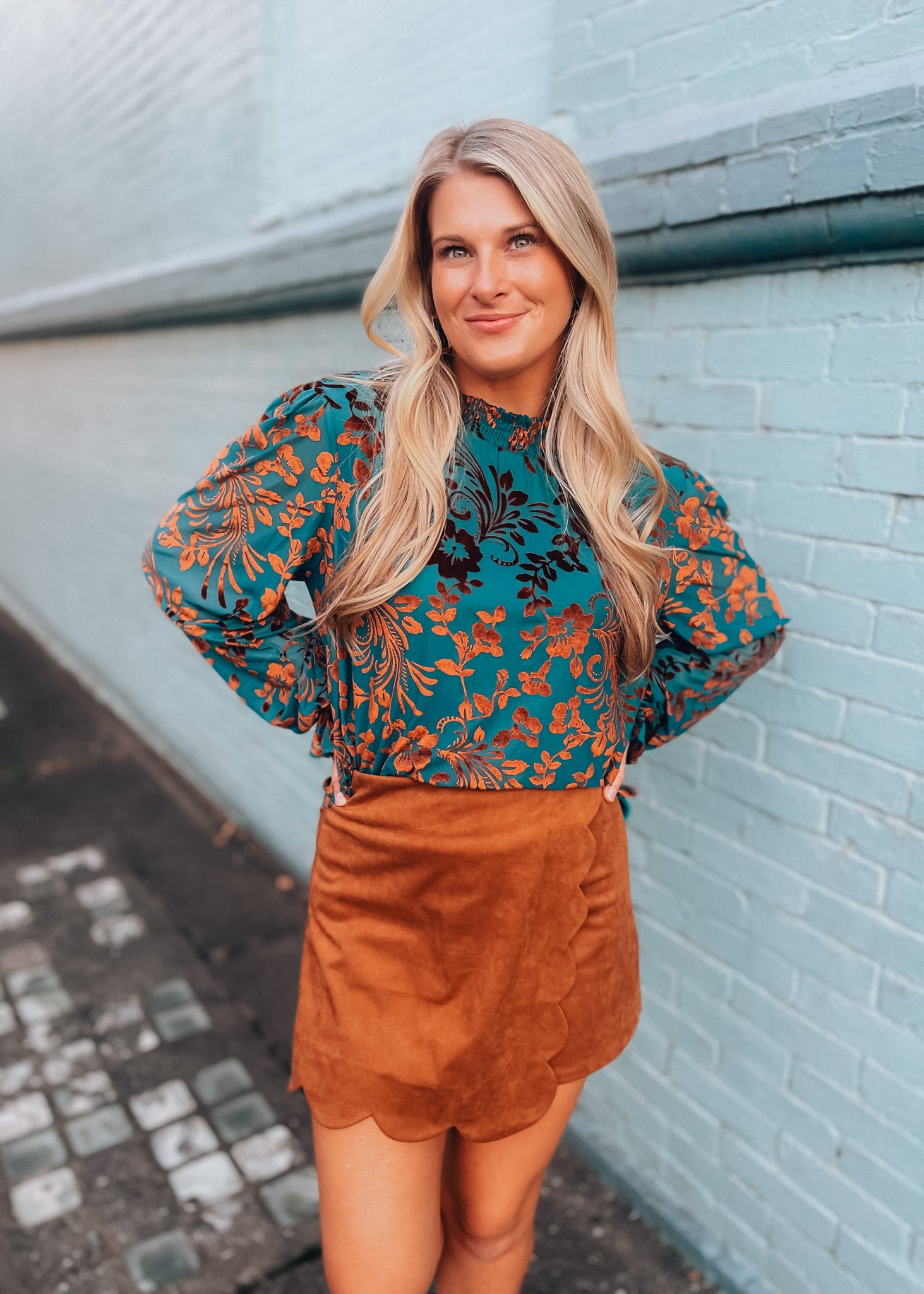 Woman wearing a floral blouse and brown skirt against a light blue wall.