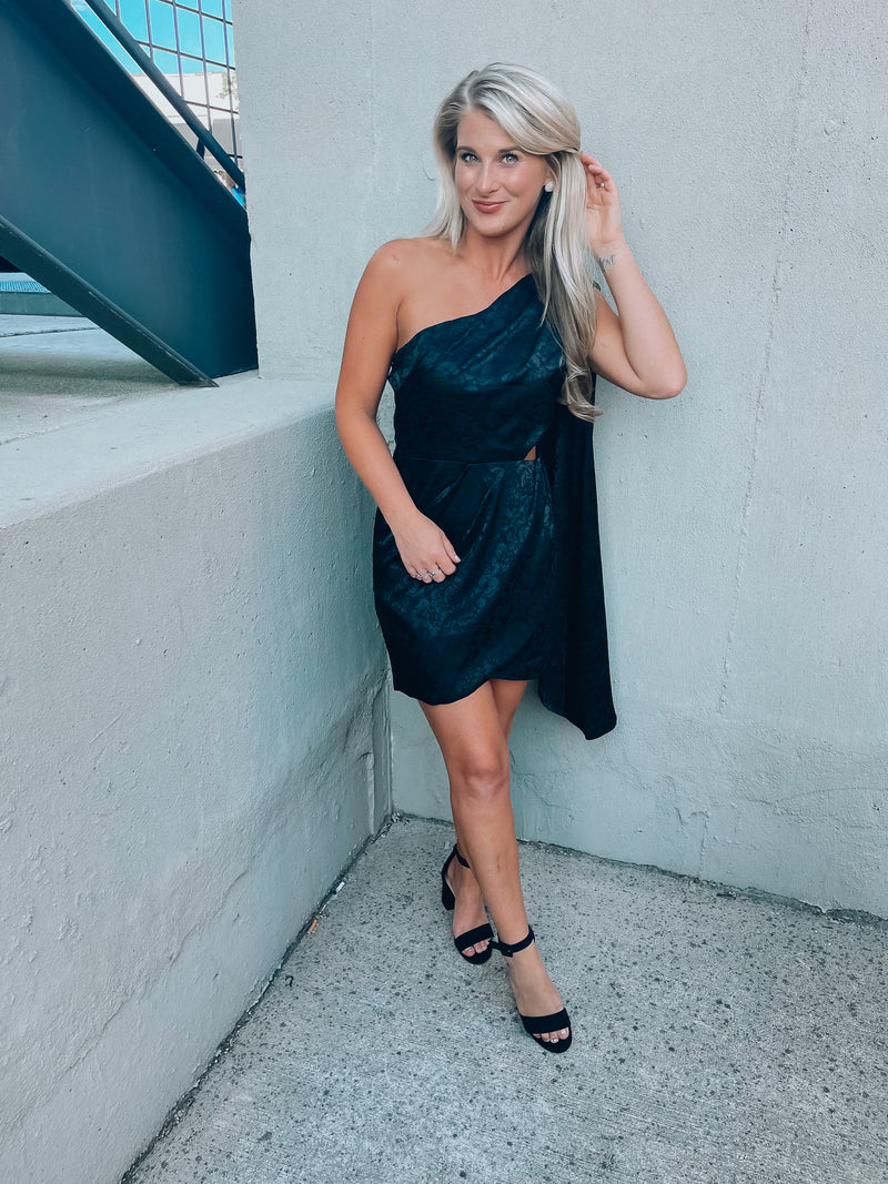 Black satin dress features subtle floral design, side cutout, with a draped, one shoulder detail. Runs true to size!  - Jacquard print - Draped one shoulder - Side ruffle detail - Pleated - Side zipper - 100% Polyester - Lined