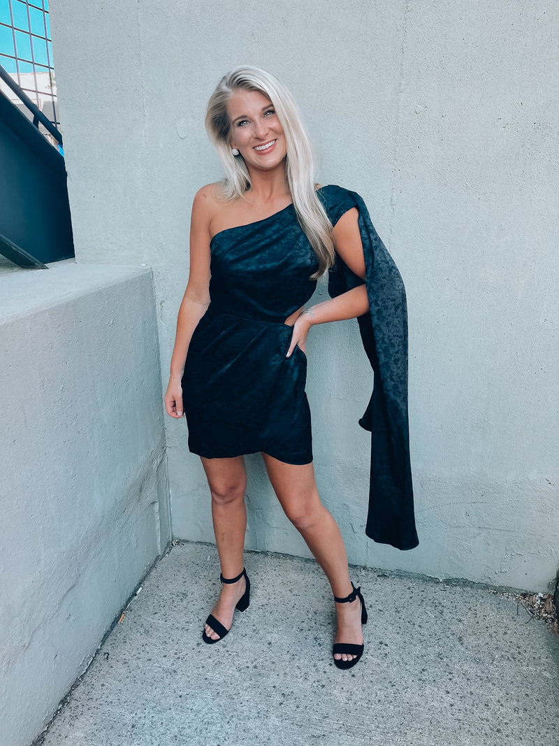 Black satin dress features subtle floral design, side cutout, with a draped, one shoulder detail. Runs true to size!  - Jacquard print - Draped one shoulder - Side ruffle detail - Pleated - Side zipper - 100% Polyester - Lined